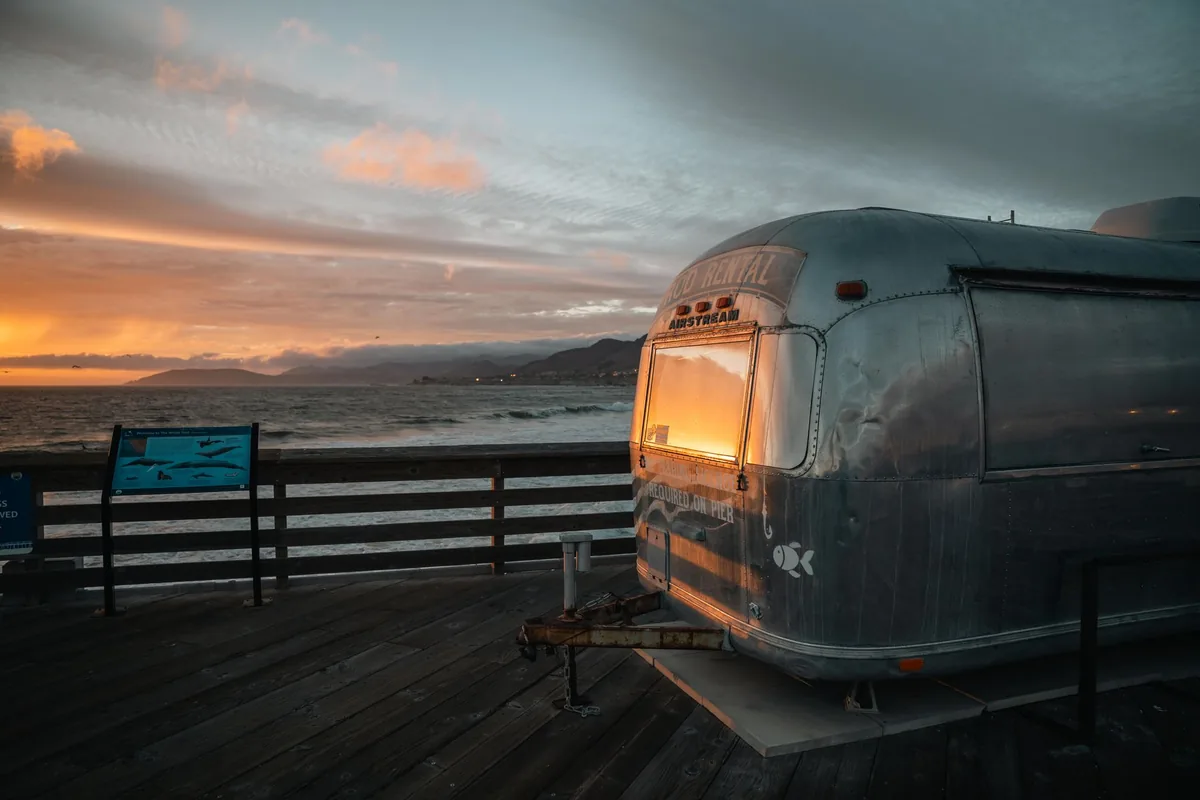 A silver travel trailer at the coast with warm light glowing from its windows at sunset