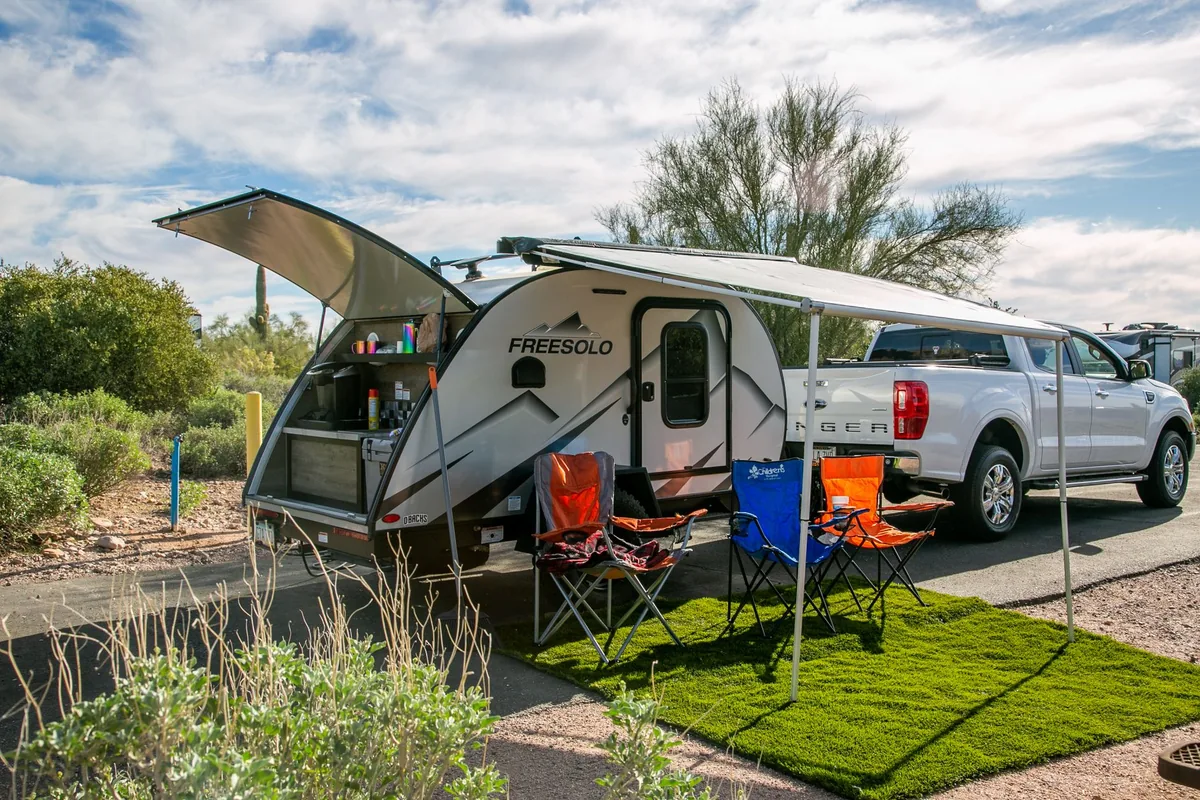 A modern silver travel trailer with awning and chairs set up beside a pickup