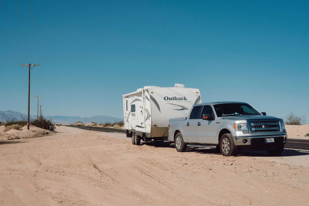 A pickup truck towing a white travel trailer on a dirt road under a clear sky